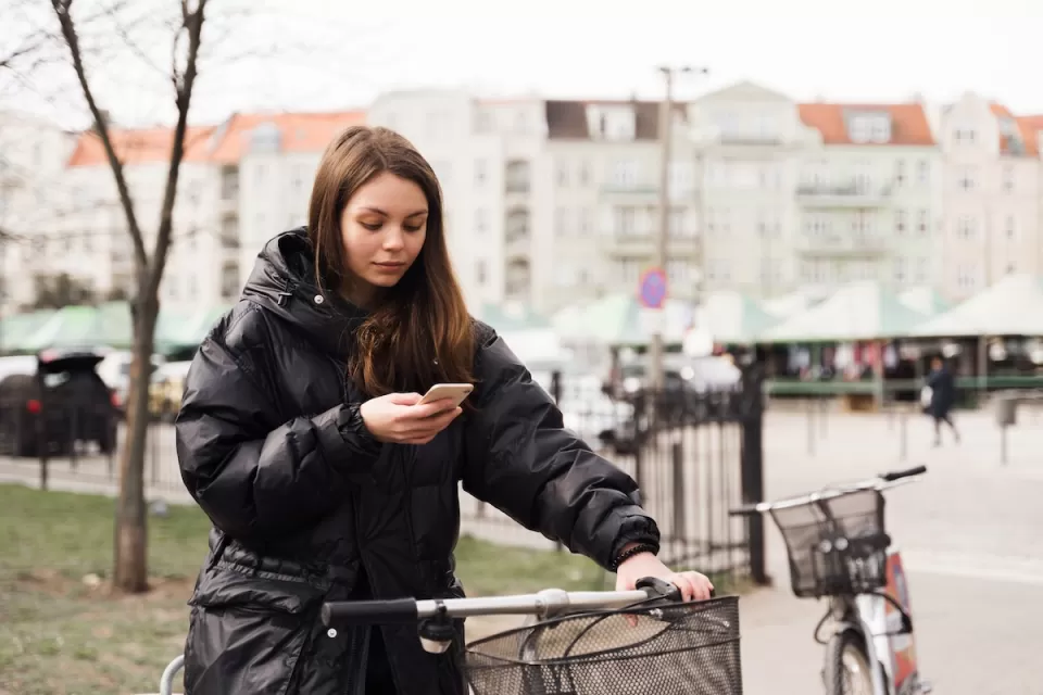 Woman stands next to a bike with one hand on handlebar holding her mobile phone.